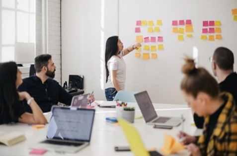 Woman pointing at sticky notes on a whiteboard while those at table watch her